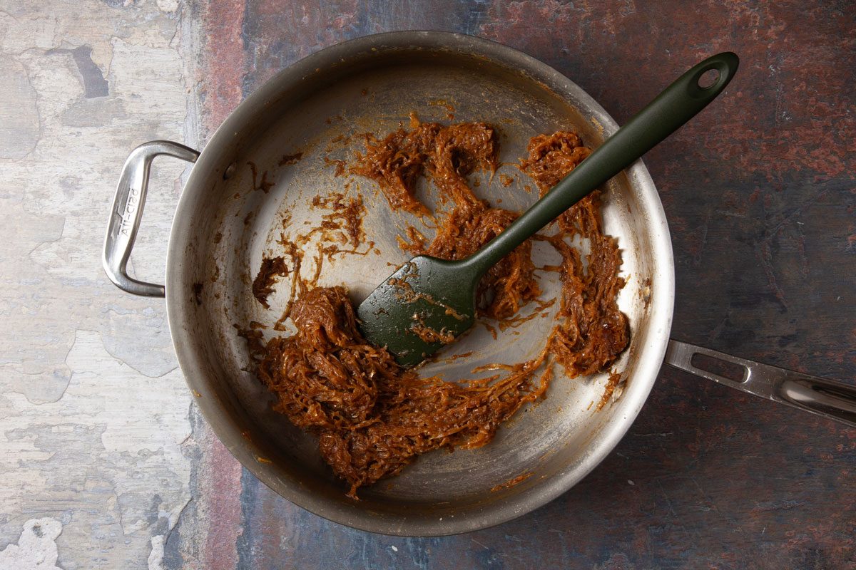 Overhead shot of a stainless steel pan containing a small amount of brown caramelized onion paste with a green spatula resting inside, placed on a mottled surface.