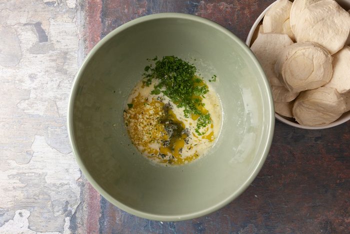 Overhead shot of a green bowl containing melted butter, chopped herbs, and seasoning, placed beside a plate of uncooked biscuit dough rounds on a textured surface.