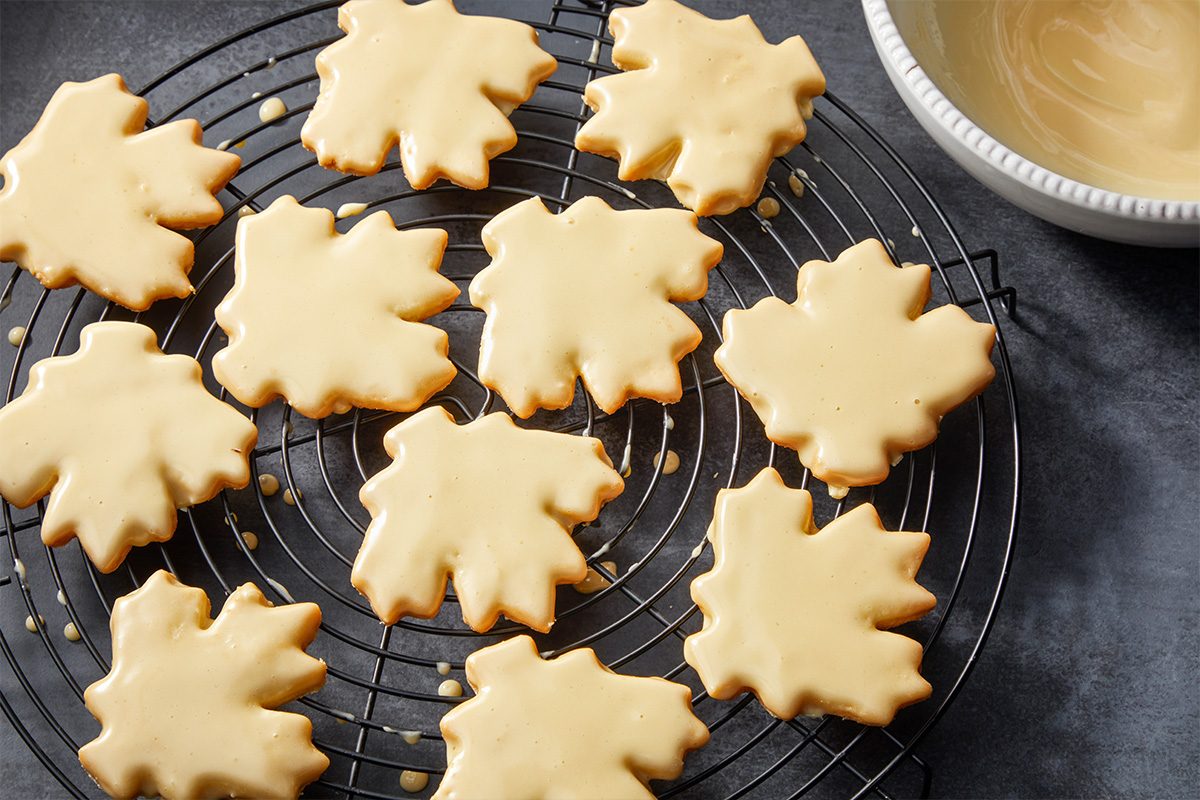 Overhead shot of Glazed Maple Shortbread Cookies with light beige icing on a round cooling rack, with a bowl of icing nearby on a dark surface;