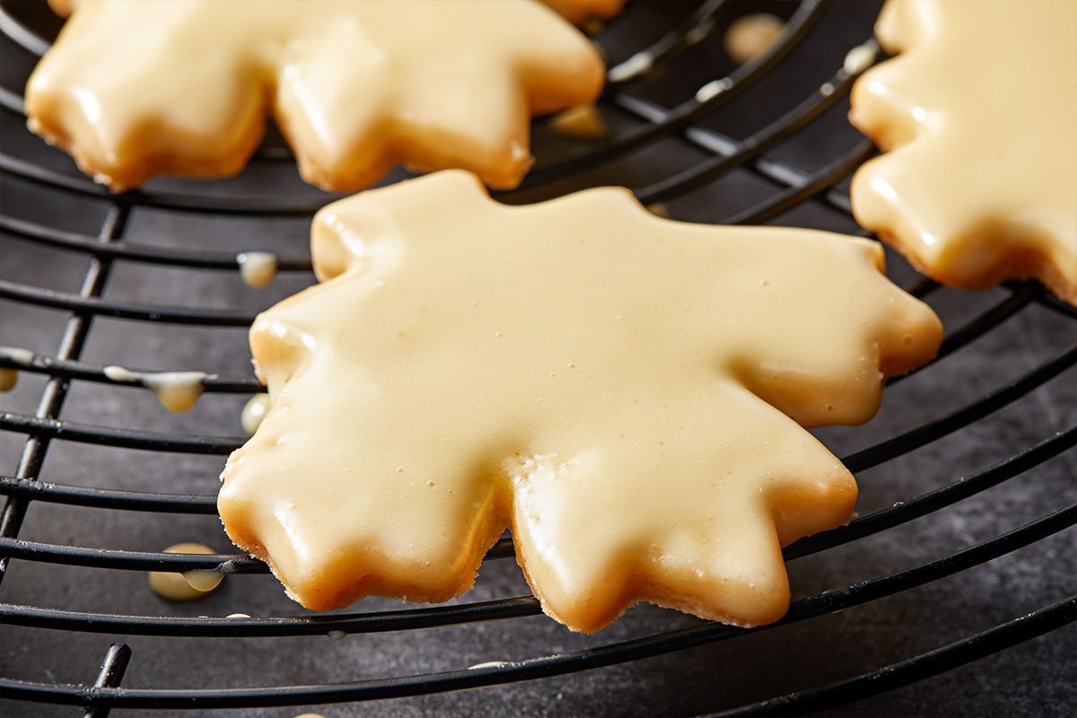 Closeup shot of a Glazed Maple Shortbread Cookie shaped like a maple leaf, coated with glossy icing on a black cooling rack, with small drips of icing nearby;