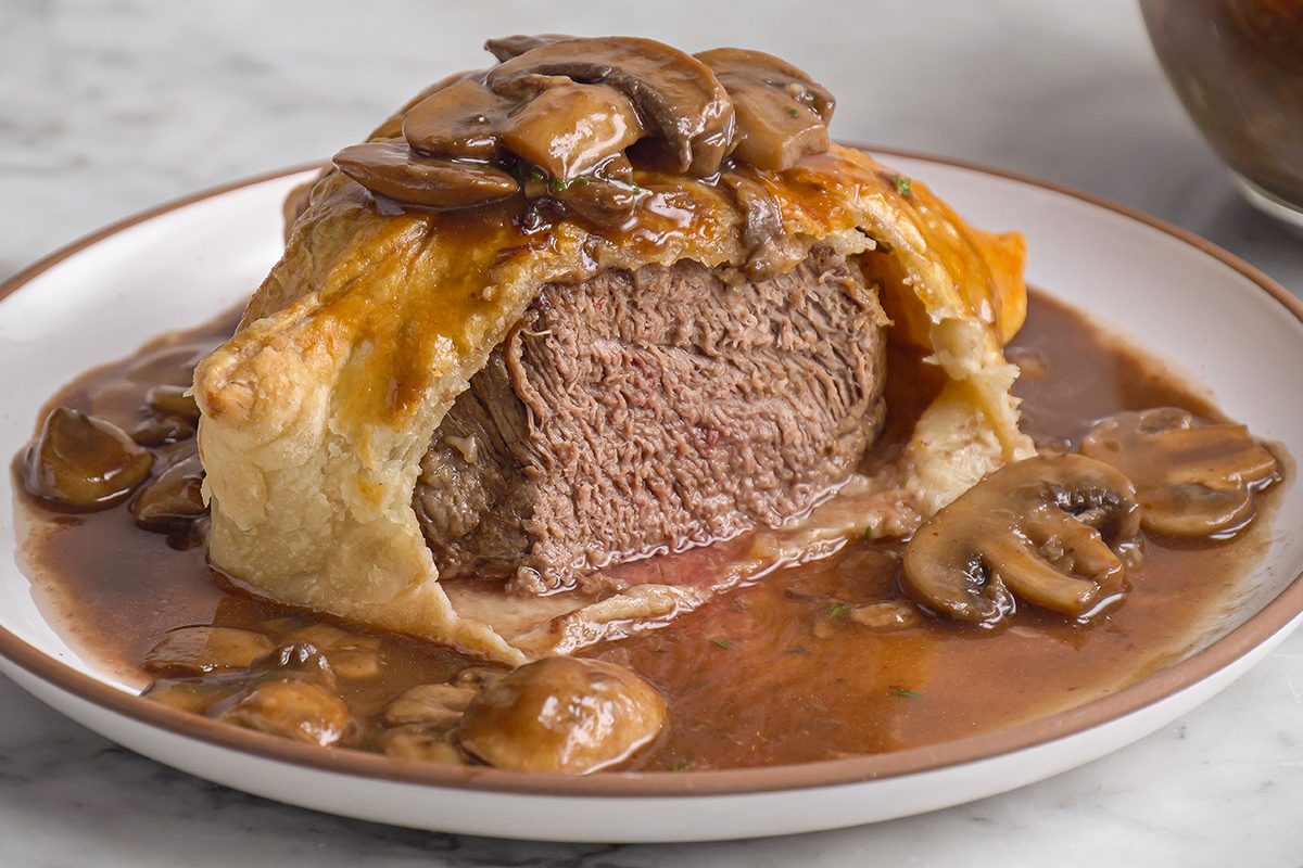 Close shot of a plate with individual Beef Wellingtons cut open, revealing medium beef inside golden pastry, served with sautéed mushrooms and mushroom sauce;