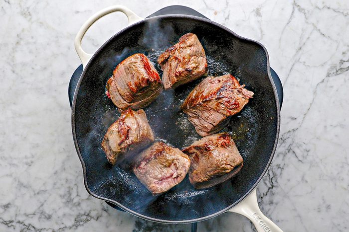 Overhead shot of six pieces of seared meat cooking in a black cast-iron skillet with a white handle, set on a white marble countertop; Steam rises from the pan;