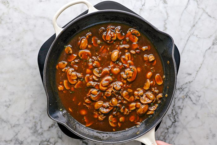Overhead shot of a skillet filled with brown mushroom sauce on a marble countertop; The sauce contains sliced mushrooms and herbs, with a hand holding the skillet handle in the lower right corner;