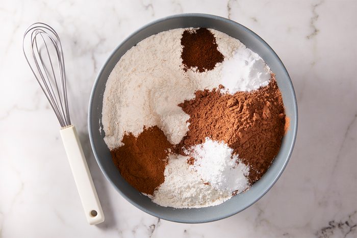 A gray bowl filled with flour, cocoa powder, baking soda, baking powder, and spices sits on a marble surface next to a metal whisk with a white handle.