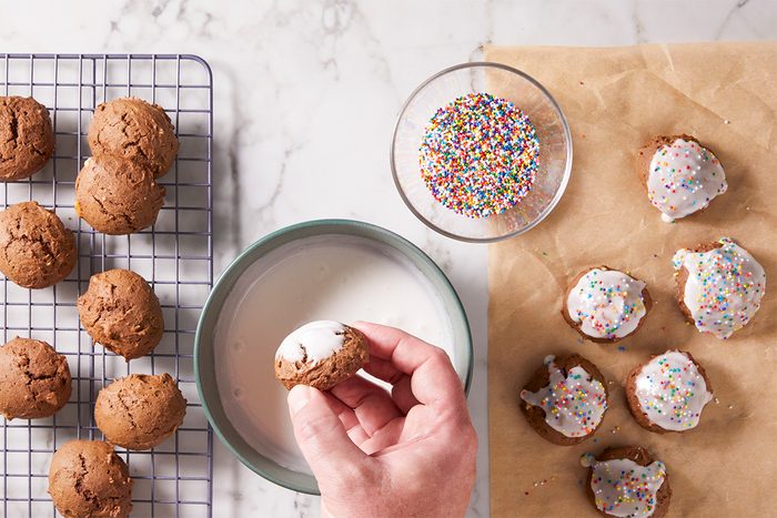 A hand dips a brown cookie into white icing. Nearby, undecorated cookies cool on a wire rack, while iced cookies with colorful sprinkles rest on parchment paper next to a bowl of rainbow sprinkles.