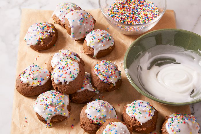 Chocolate cookies with white icing and colorful sprinkles are arranged on parchment paper. Nearby, there is a bowl of icing and a bowl of rainbow sprinkles.