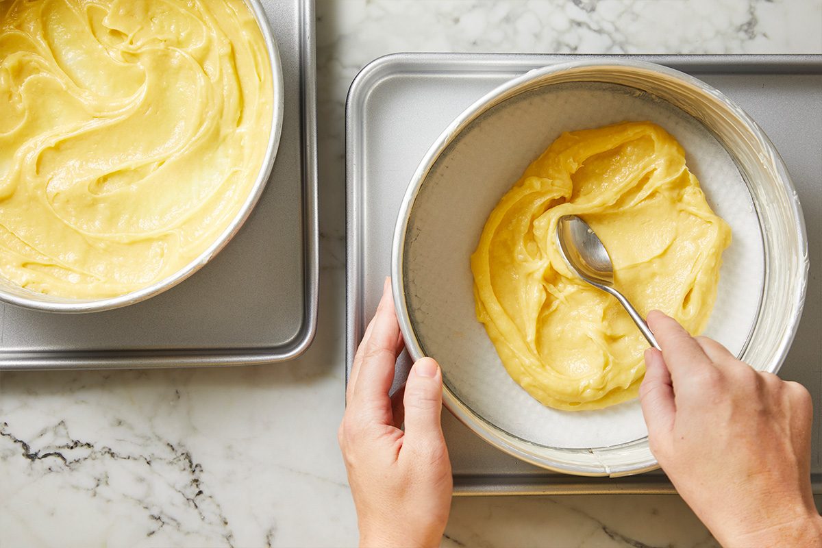 Overhead shot of a person spreading yellow cake batter in a round cake pan lined with parchment paper using a spoon, with another pan of batter nearby on a marble countertop;