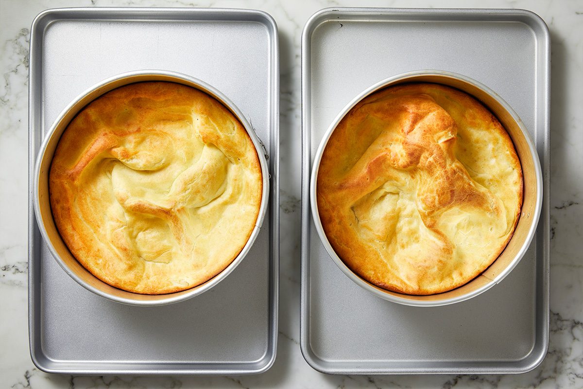 Overhead shot of two round, golden-brown Dutch baby pancakes in cake pans, side by side on a marble countertop, with puffed, uneven surfaces and lightly browned edges; 