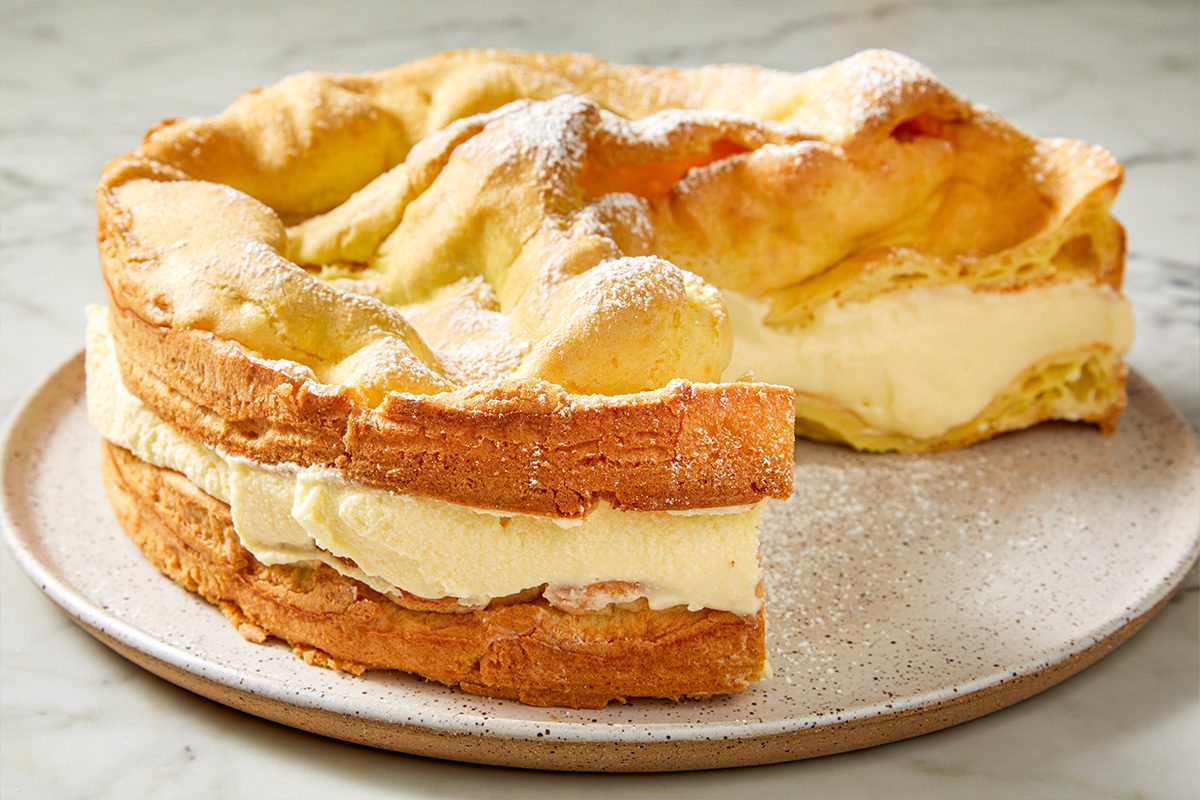 3/4 angle view shot of a slice of Karpatka Cake with creamy filling and a golden crust, dusted with powdered sugar, sitting on a plate on a marble surface;