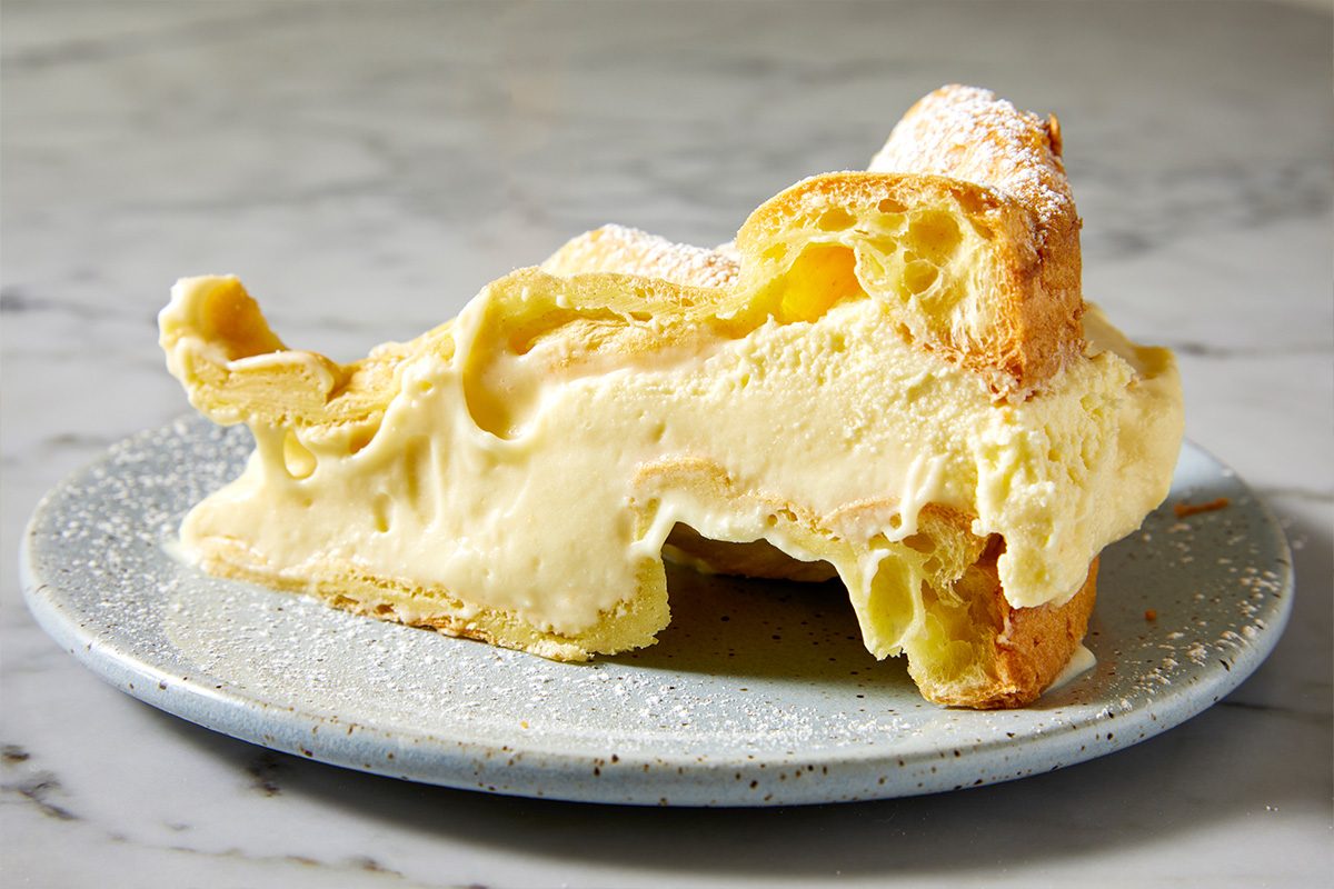 Closeup shot of a slice of Karpatka Cake on a speckled plate, featuring golden pastry, creamy filling, and a dusting of powdered sugar on top;