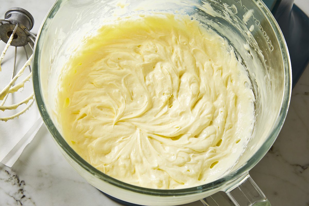 Overhead shot of a glass mixing bowl filled with smooth, creamy batter on a marble countertop, next to a metal hand mixer with batter on its beaters and a white cloth;