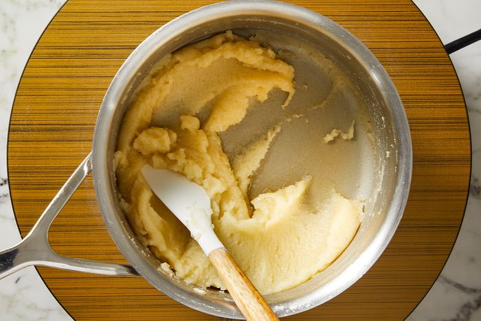 Overhead shot of a saucepan containing thick, pale dough being mixed with a white spatula with a wooden handle, placed on a round wooden surface;