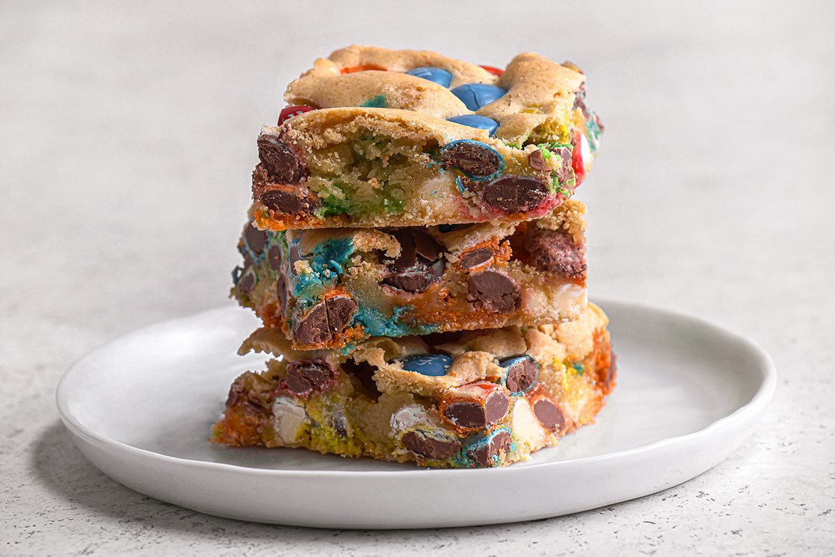 A stack of three colorful candy and chocolate chip blondies sits on a white plate, set against a light, neutral background.