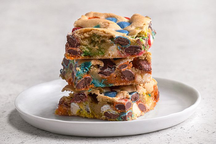 A stack of three colorful candy and chocolate chip blondies sits on a white plate, set against a light, neutral background.