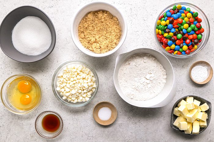 A top-down view of baking ingredients in bowls: sugar, crushed graham crackers, colorful candy-coated chocolates, eggs, white chocolate chips, flour, butter, vanilla extract, salt, and baking powder on a light surface.