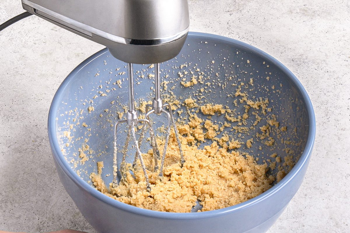 A hand mixer is blending cookie dough in a large blue mixing bowl on a light-colored countertop.