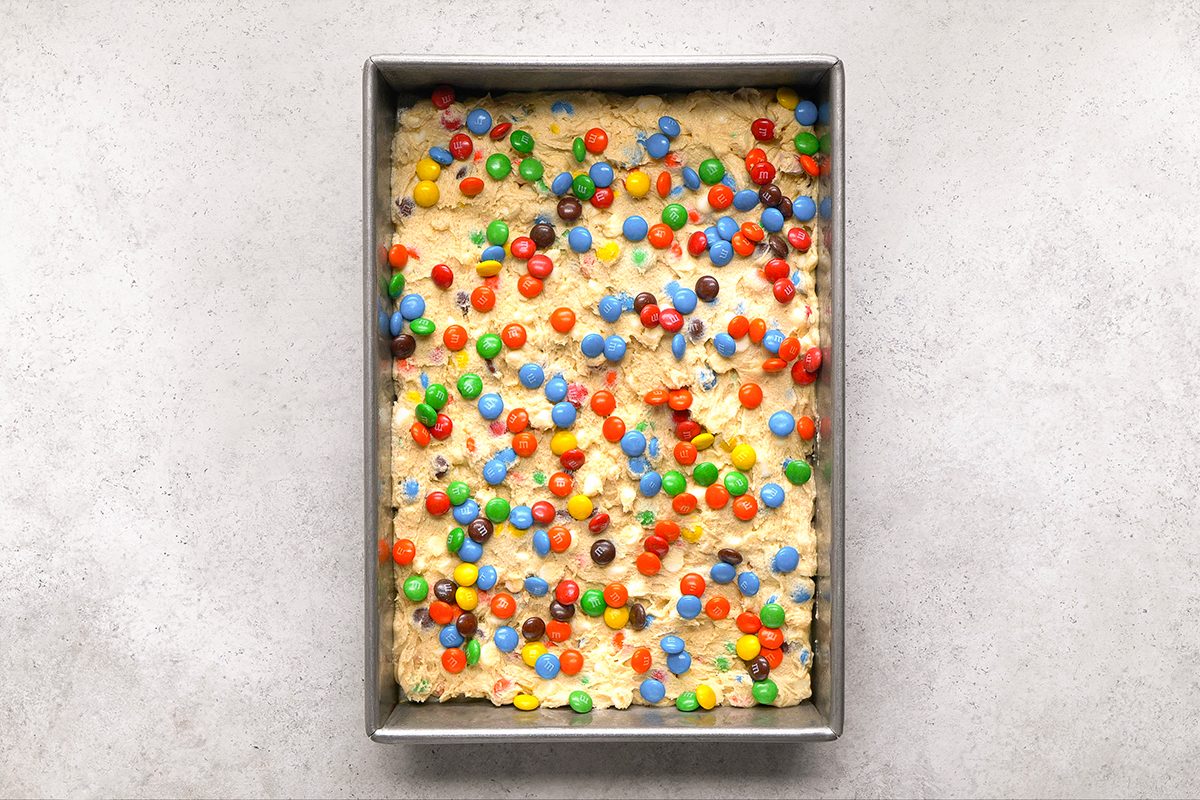 A rectangular baking pan filled with unbaked cookie dough topped with colorful candy-coated chocolates, sitting on a light gray surface.