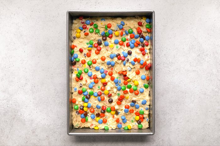 A rectangular baking pan filled with unbaked cookie dough topped with colorful candy-coated chocolates, sitting on a light gray surface.