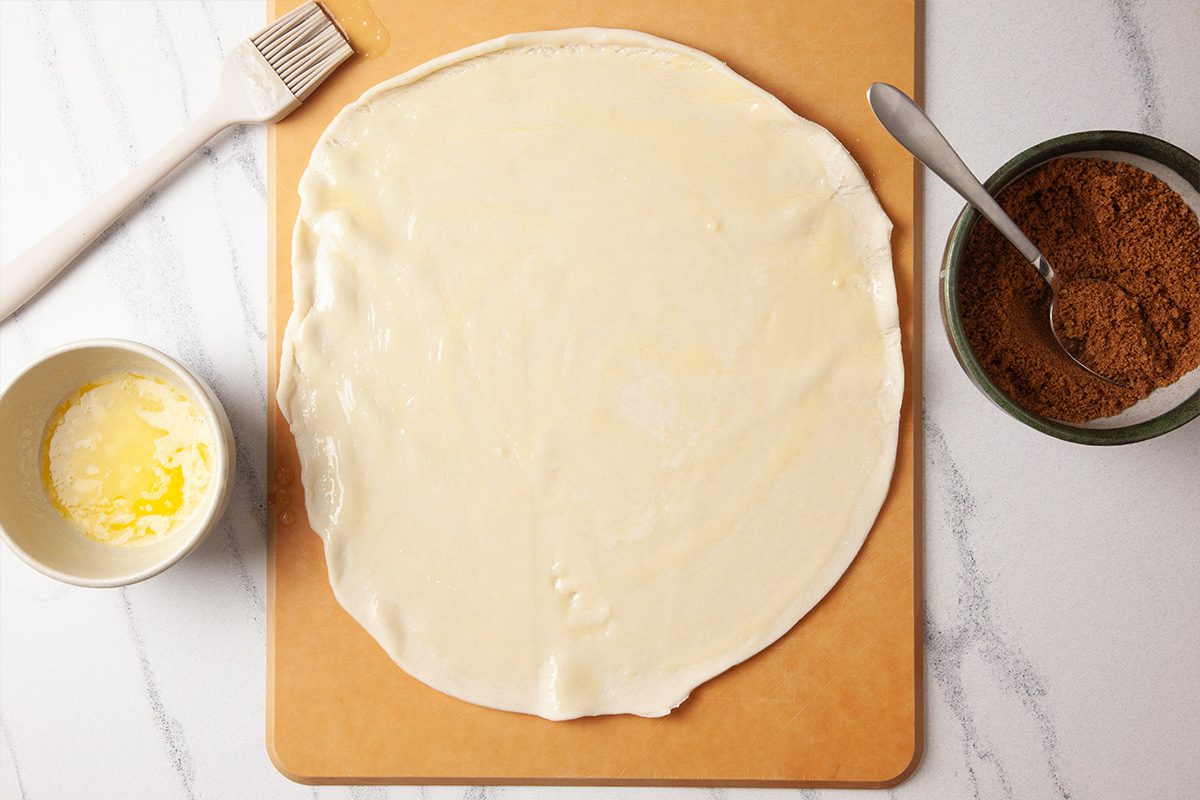 Overhead shot of a round sheet of rolled-out dough brushed with melted butter on a cutting board, with a pastry brush, a bowl of melted butter, a spoon, and a bowl of brown sugar mixture nearby;