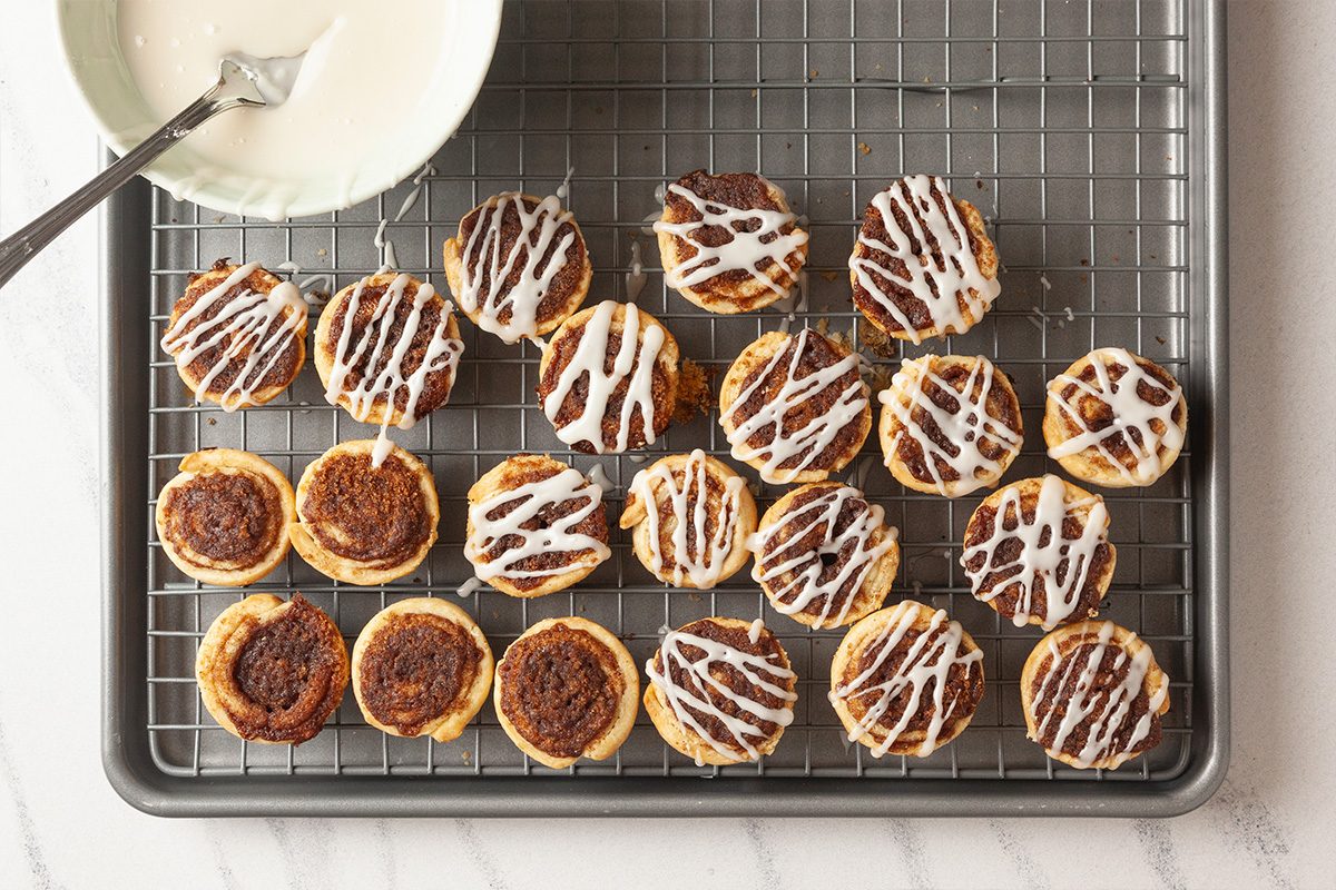 Overhead shot of small cinnamon rolls on a cooling rack, some drizzled with white icing. A bowl of extra icing with a spoon sits nearby