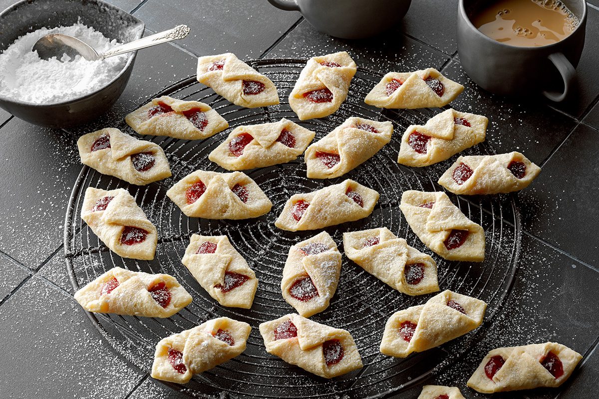 A cooling rack holds dozens of diamond-shaped cookies filled with red jam and dusted with powdered sugar. A bowl of powdered sugar with a spoon and two cups of coffee are nearby on a dark tile surface.
