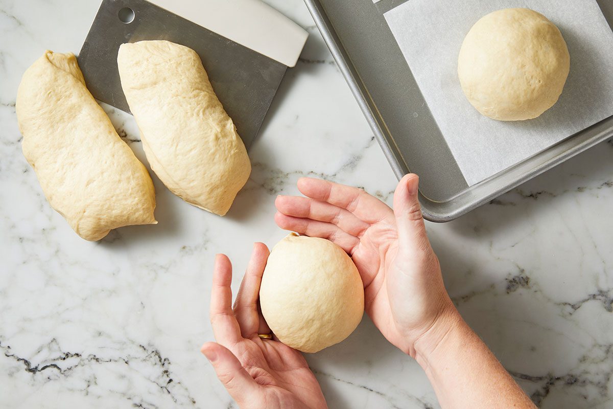 Overhead shot of hands shaping a ball of dough on a marble surface, with two dough pieces, a bench scraper, and a parchment-lined baking sheet holding another dough ball nearby.