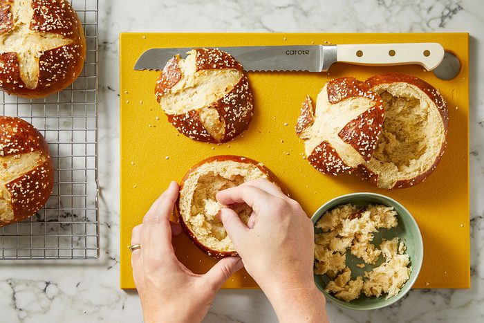 Overhead shot of two hands hollowing out a pretzel bread bowl on a yellow board, with additional bowls, a knife, and a green bowl of crumbs nearby on a marble surface.