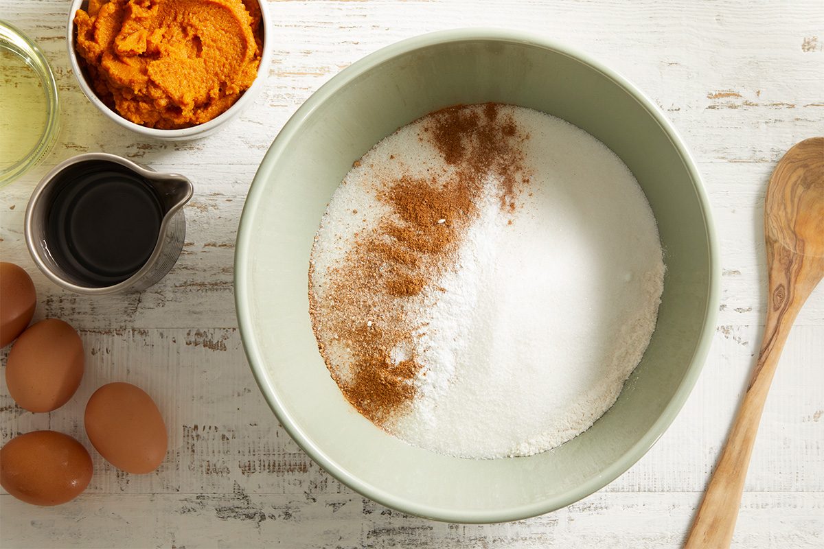 A bowl with sugar and spices sits on a white wooden surface, surrounded by eggs, a wooden spoon, a cup of dark liquid, oil, and a bowl of orange puree.
