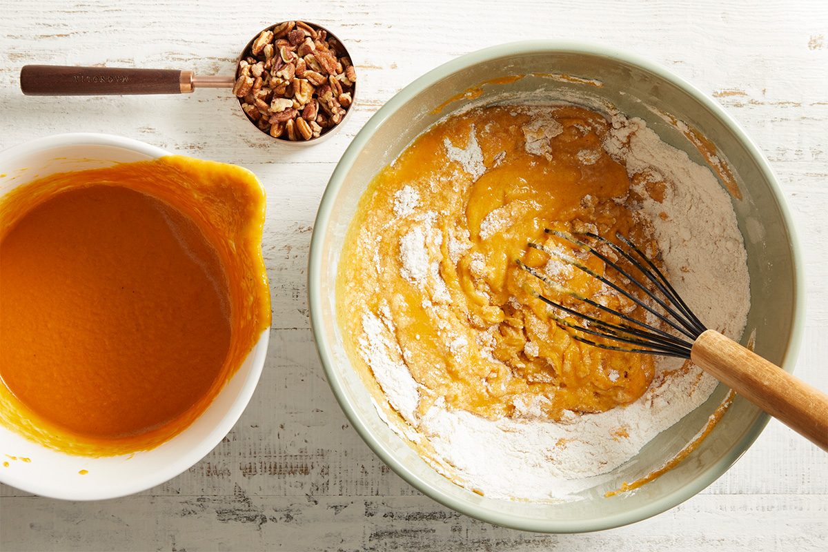 A bowl of orange pumpkin puree, a bowl with flour and pumpkin mix being whisked, and a measuring cup of chopped pecans are arranged on a white surface, ready for baking.
