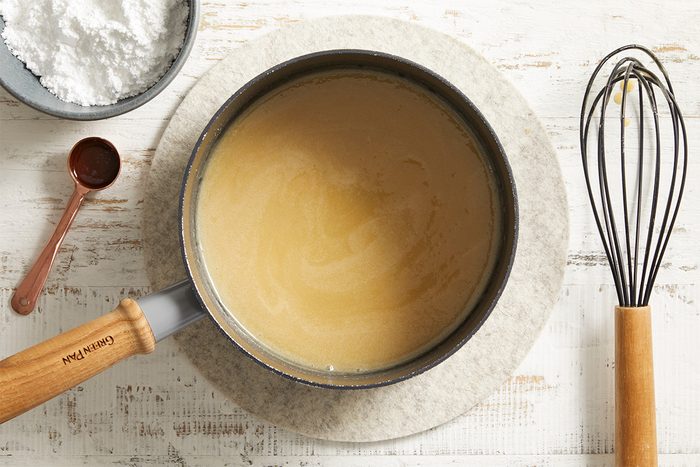 A saucepan with a light brown sauce sits on a round trivet. Nearby are a metal whisk with a wooden handle, a spoon with brown liquid, and a bowl of white powdered ingredient, all on a rustic white table.