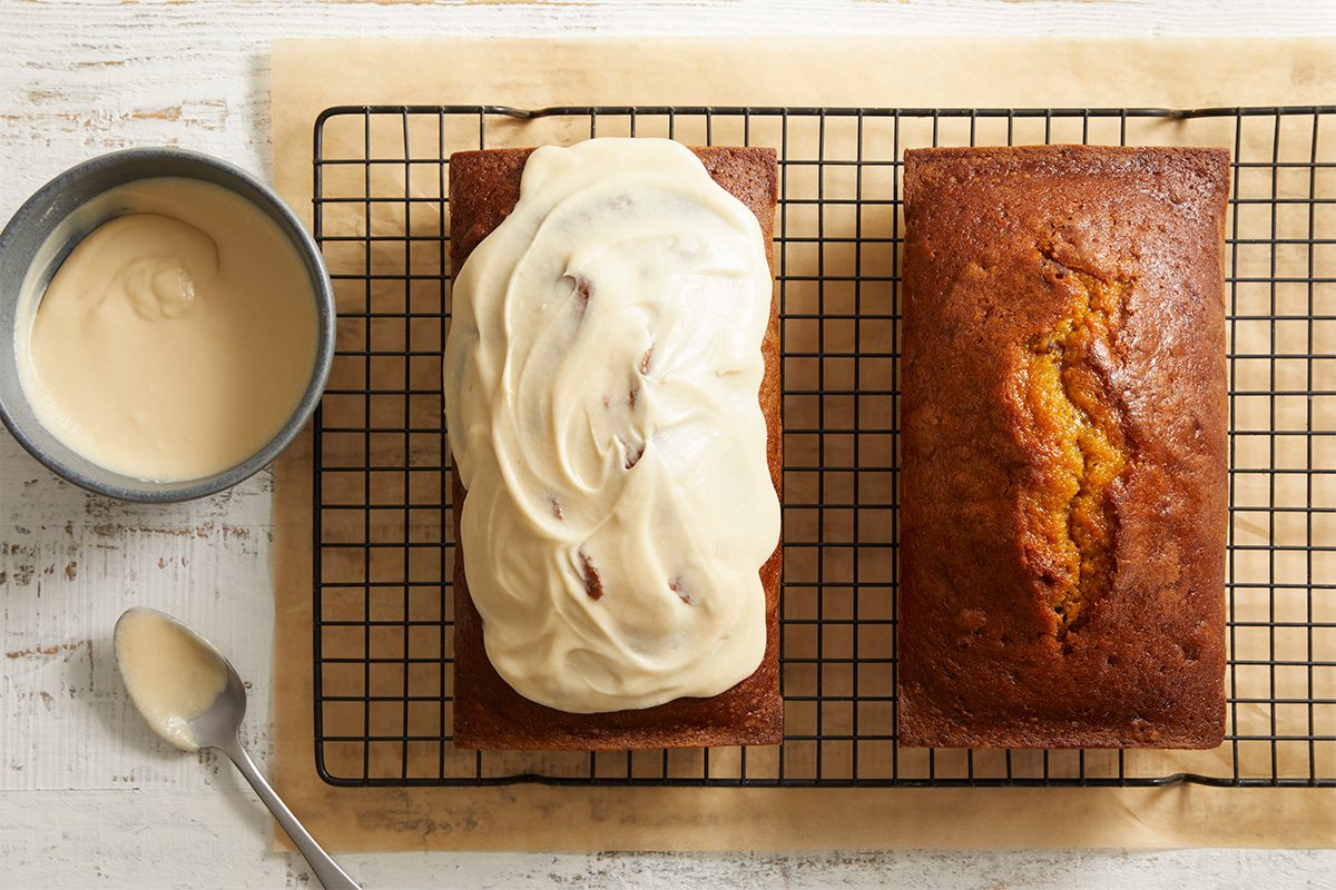 Two rectangular loaves of bread sit on a cooling rack; one loaf is plain, the other is topped with creamy frosting. 