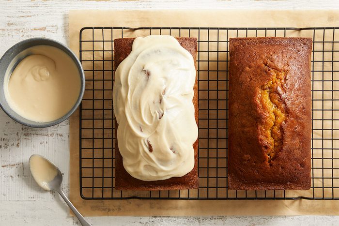 Two rectangular loaves of bread sit on a cooling rack; one loaf is plain, the other is topped with creamy frosting.