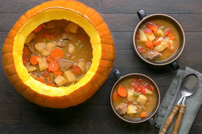 A large hollowed pumpkin filled with hearty stew containing beef, potatoes, carrots, and tomatoes, sits on a dark wooden table beside two bowls of the same stew, with a gray napkin and utensils next to them.