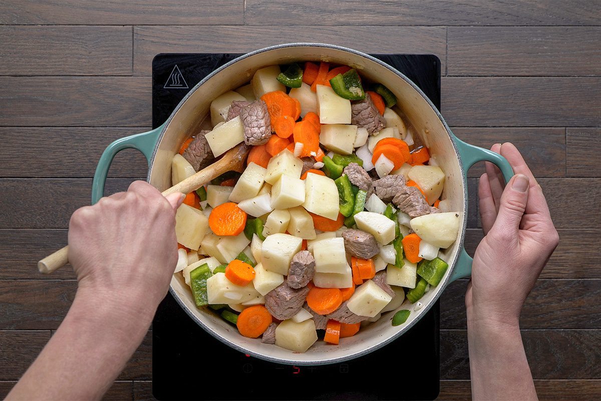 A person stirs a pot filled with chunks of beef, potatoes, carrots, and green bell peppers on a stovetop. The ingredients are colorful and uncooked.