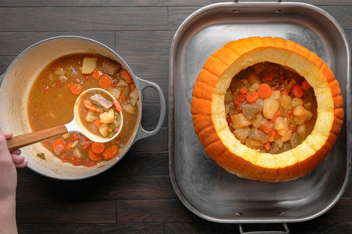 A hand ladles vegetable stew with carrots and potatoes from a pot into a hollowed-out pumpkin sitting in a large roasting pan on a dark wooden surface.