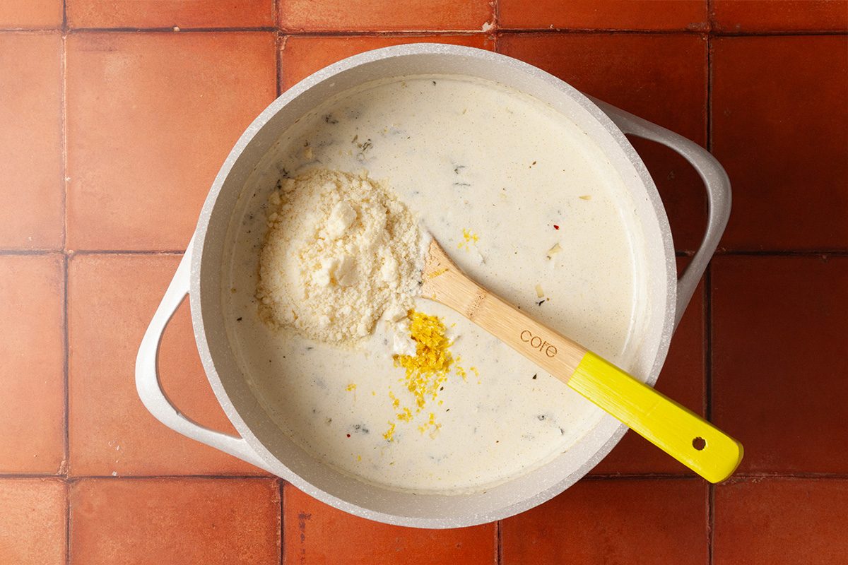 Overhead shot of a white pot of creamy soup on a tiled surface, with a wooden spoon featuring a yellow handle holding grated cheese and lemon zest over the soup, ready to be mixed in;