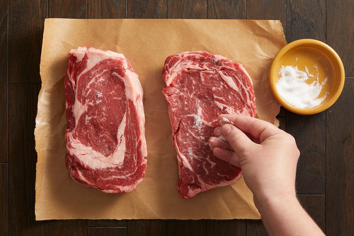 Overhead shot of two raw ribeye Steaks Alfredo being seasoned on brown parchment paper with a small bowl of salt nearby.