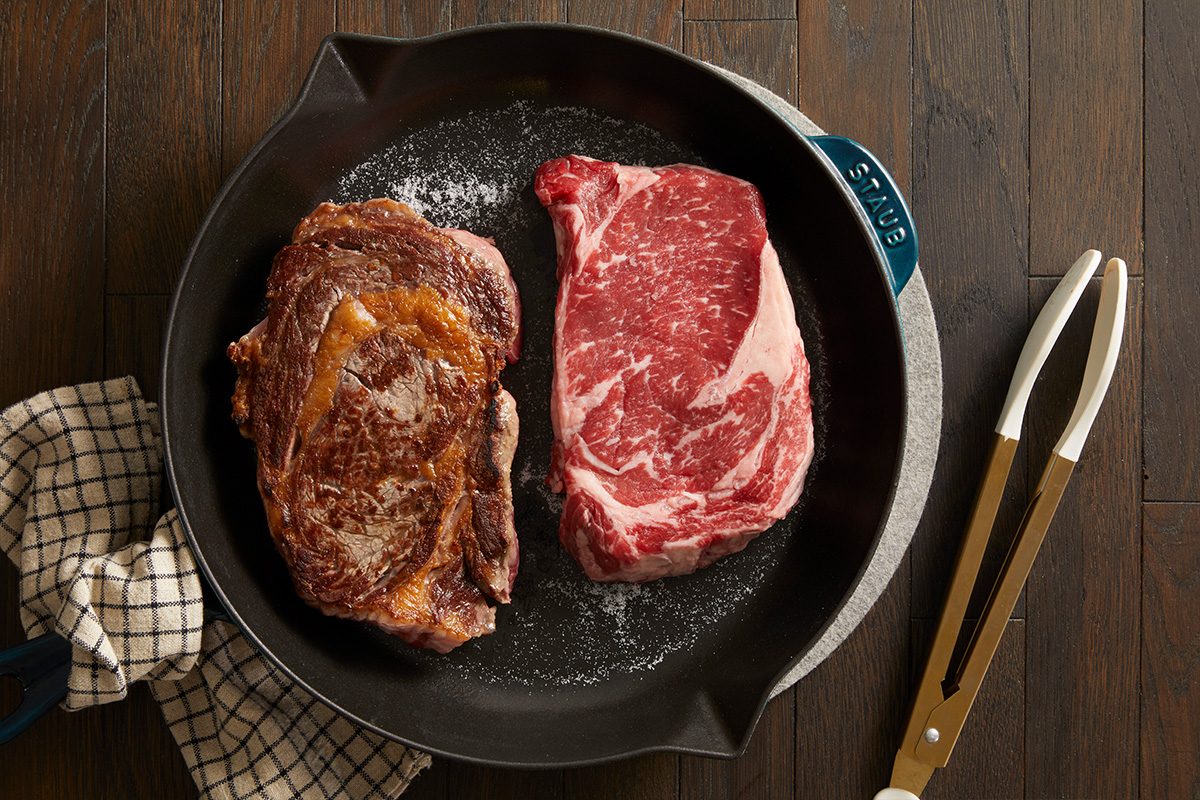 Overhead shot of two ribeye steaks searing in a black skillet on a dark wooden surface with metal tongs nearby