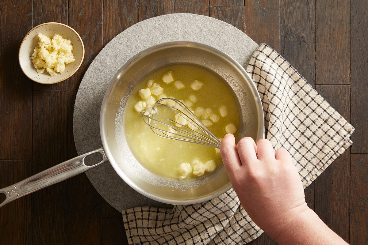 Overhead shot of butter melting in a saucepan with minced garlic being whisked in, on a wooden background with a checkered cloth.