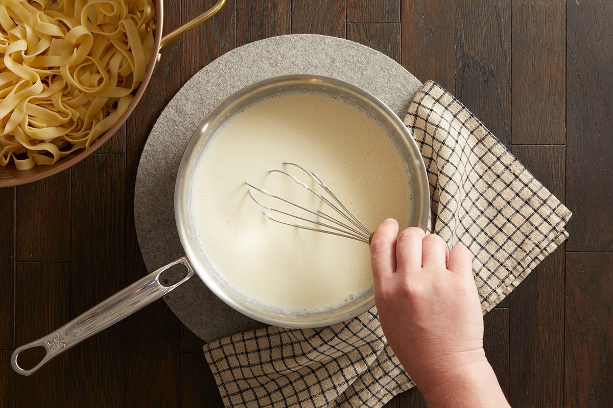 Overhead shot of creamy Alfredo sauce being whisked in a saucepan on a wooden surface, next to a bowl of uncooked fettuccine.