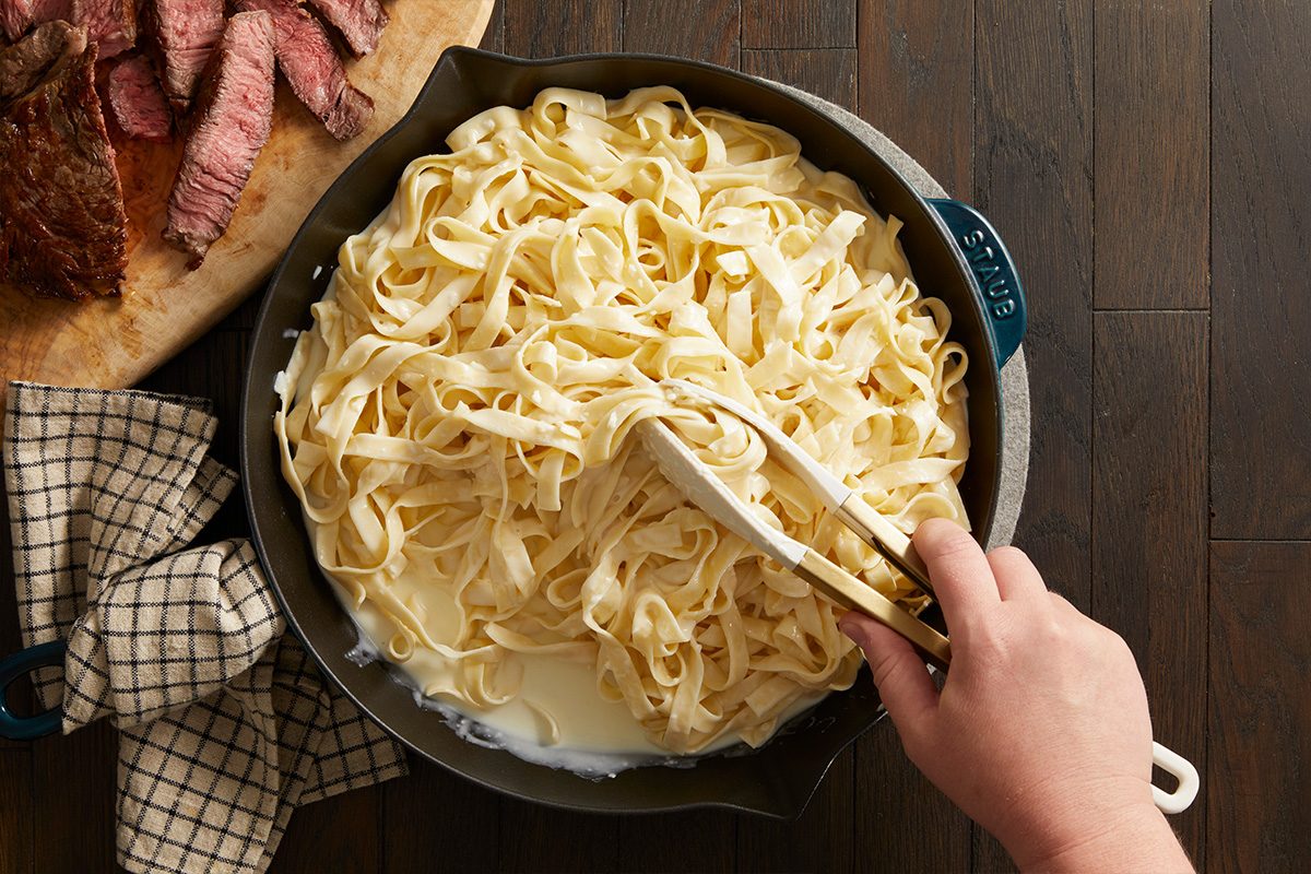 Overhead shot of cooked pasta being tossed in Alfredo sauce in a pan with a serving spoon on a dark wooden background.