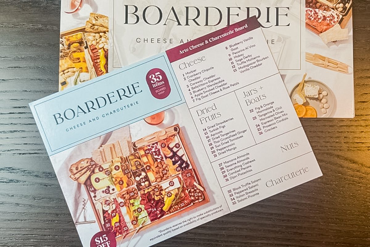 A Boarderie cheese and charcuterie kit sits on a wooden table, showing a photo of assorted meats, cheeses, and snacks on the packaging, along with a menu and ordering details.