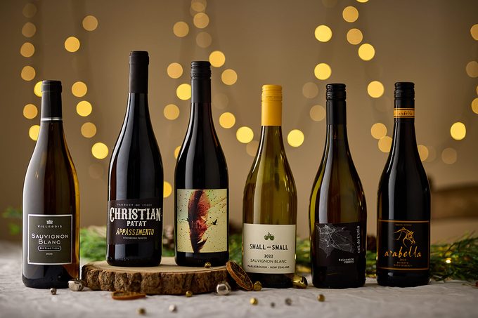 Six assorted wine bottles are arranged in a row on a table with festive string lights softly blurred in the background, creating a warm, celebratory atmosphere.