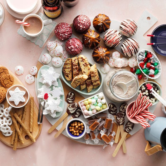 A festive assortment of holiday treats on a table, including cupcakes, cookies, biscotti, pretzels, candies, hot chocolate, and candy canes arranged on serving boards with mugs nearby.
