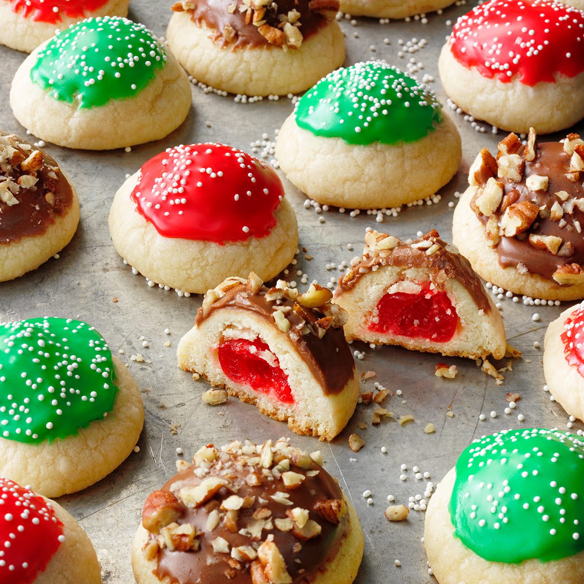 Colorful cookies topped with red, green, or chocolate icing and sprinkles. Some cookies are cut open, revealing a cherry filling inside. The cookies are arranged on a baking sheet.
