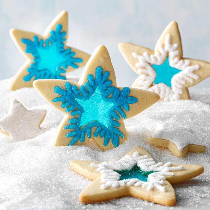 Star-shaped sugar cookies decorated with blue and white icing, arranged on a snowy, sparkling surface. Some cookies have intricate snowflake designs and translucent centers.