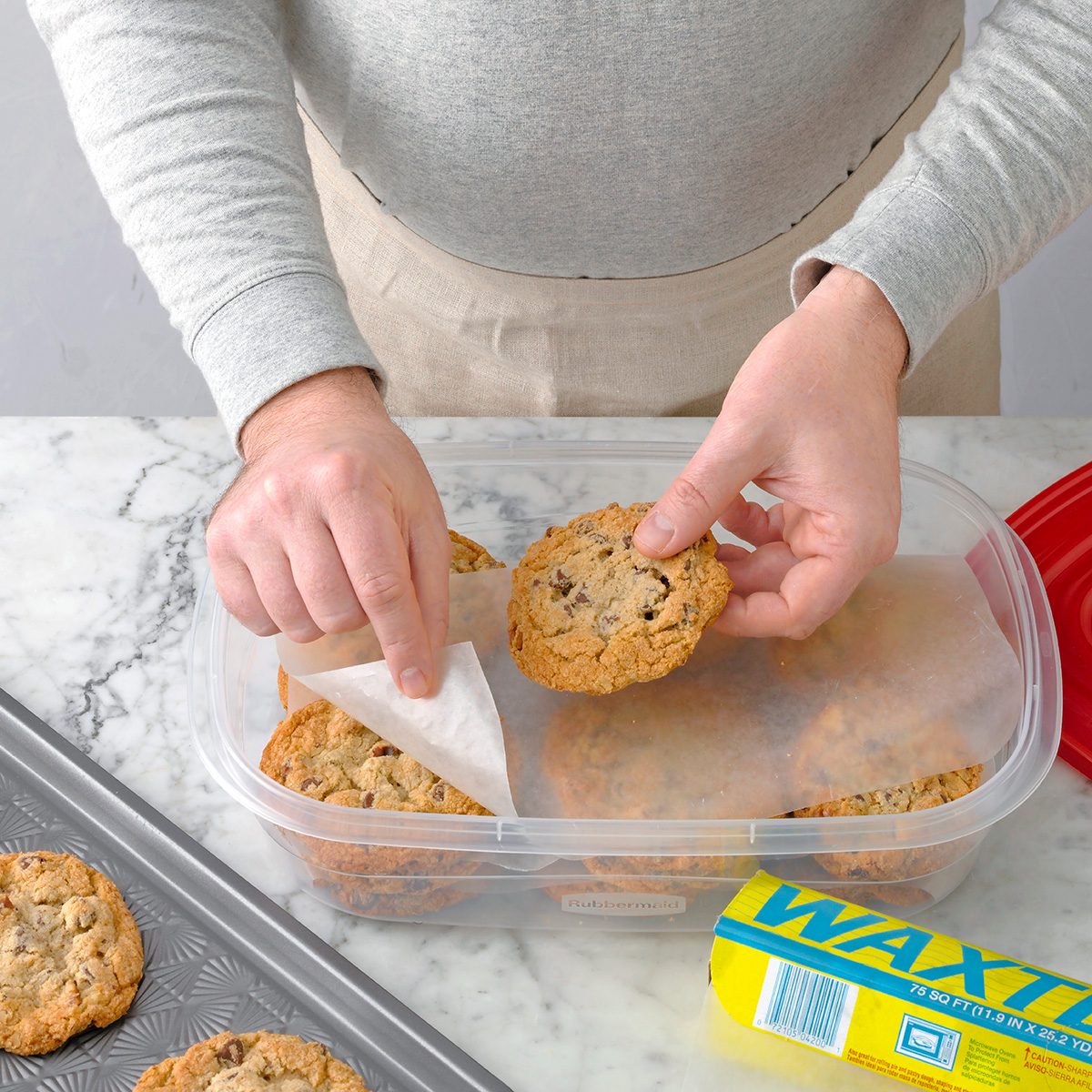 A person places chocolate chip cookies into a plastic container, separating them with sheets of wax paper on a marble countertop. A tray of cookies and a box of wax paper are nearby.