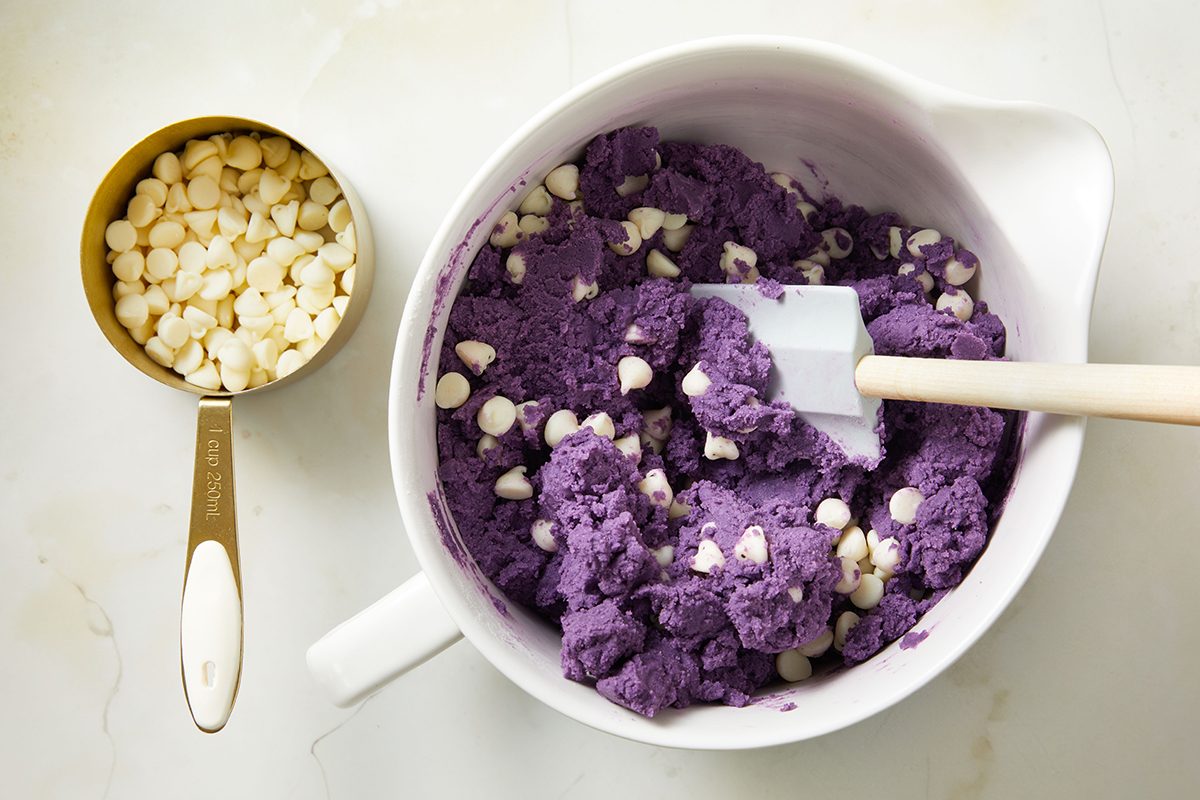 A white mixing bowl filled with vibrant purple cookie dough mixed with white chocolate chips. Beside it, a gold and white measuring cup holds extra white chocolate chips. A spatula rests in the bowl.