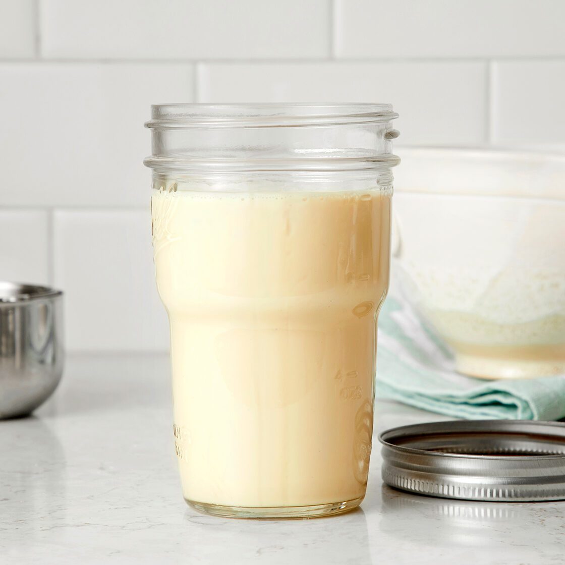 A glass jar filled with pale yellow liquid sits on a white countertop, next to a metal measuring cup, a jar lid, and a bowl with a blue cloth. White subway tiles are in the background.