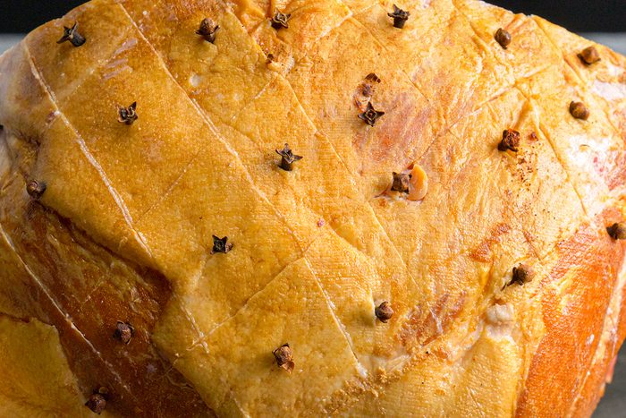 Close-up of a baked ham with a golden-brown surface, scored in a diamond pattern and studded with whole cloves at each intersection.